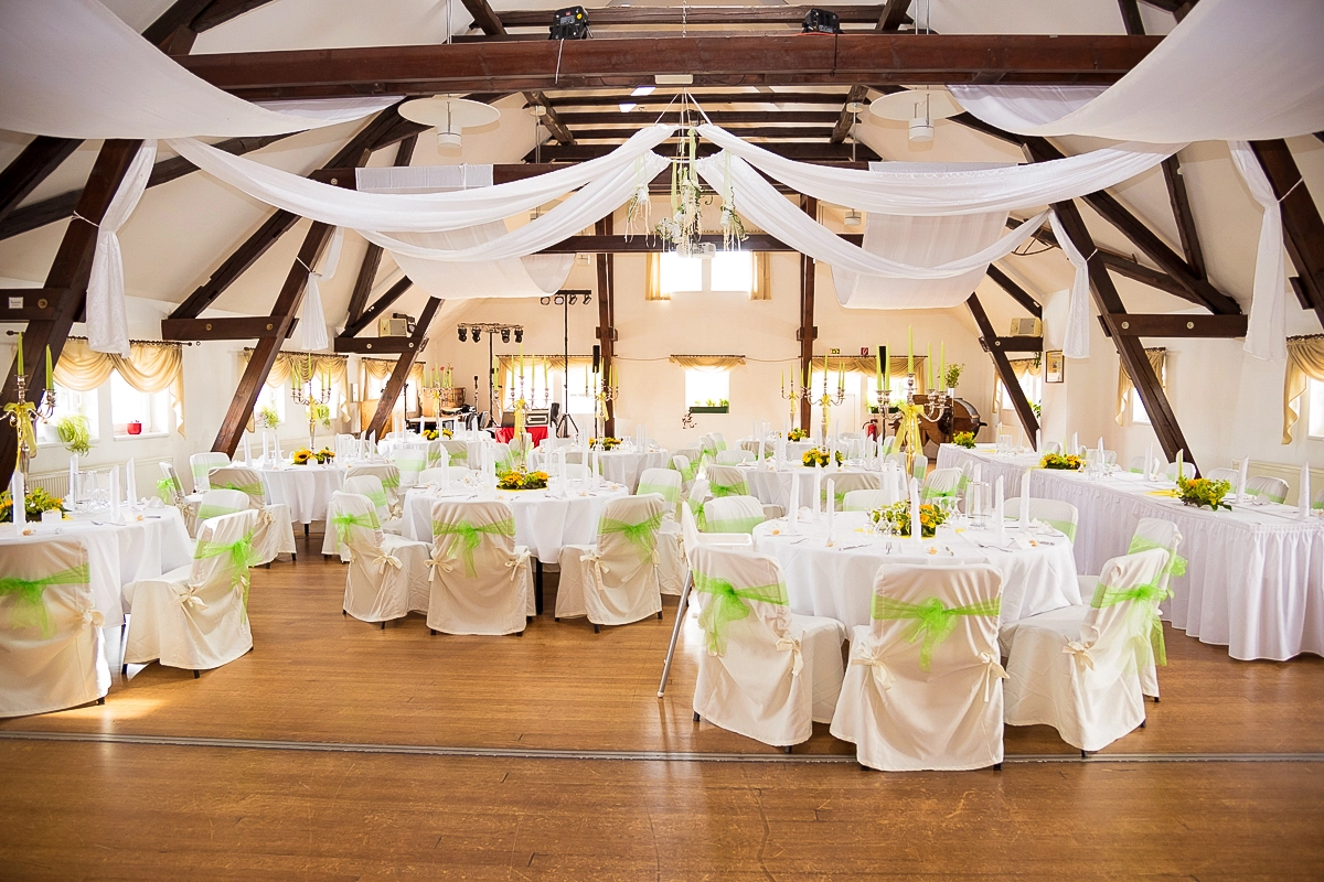 Charmante Hochzeitslocation: Dekorierter Festsaal in Thüringen Innenansicht eines grossen Hochzeitssaals mit Holzgebälk, festlich dekoriert mit weissen Stoffbahnen an der Decke. Zahlreiche runde Tische sind mit weissen Tischtüchern, Stuhlhussen mit grünen Bändern und gelben Blumendekorationen für die Feier gedeckt. Fotografiert von Michael Schalansky.
